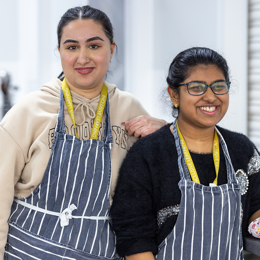 Two Female Uxbridge College students from Inclusive Learning in the kitchen at Hayes Campus. They are both wearing blue with white stripe aprons with yellow lanyards. 