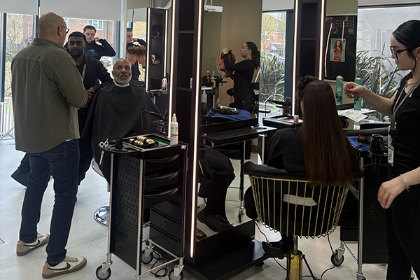 Meet the Principal Video behind the scenes footage - Clive is in the salon at Hayes campus, sitting in front of a mirror with the black cape to catch hair draped around him. He is talking to a film crew and there are other students practising hairdressing.