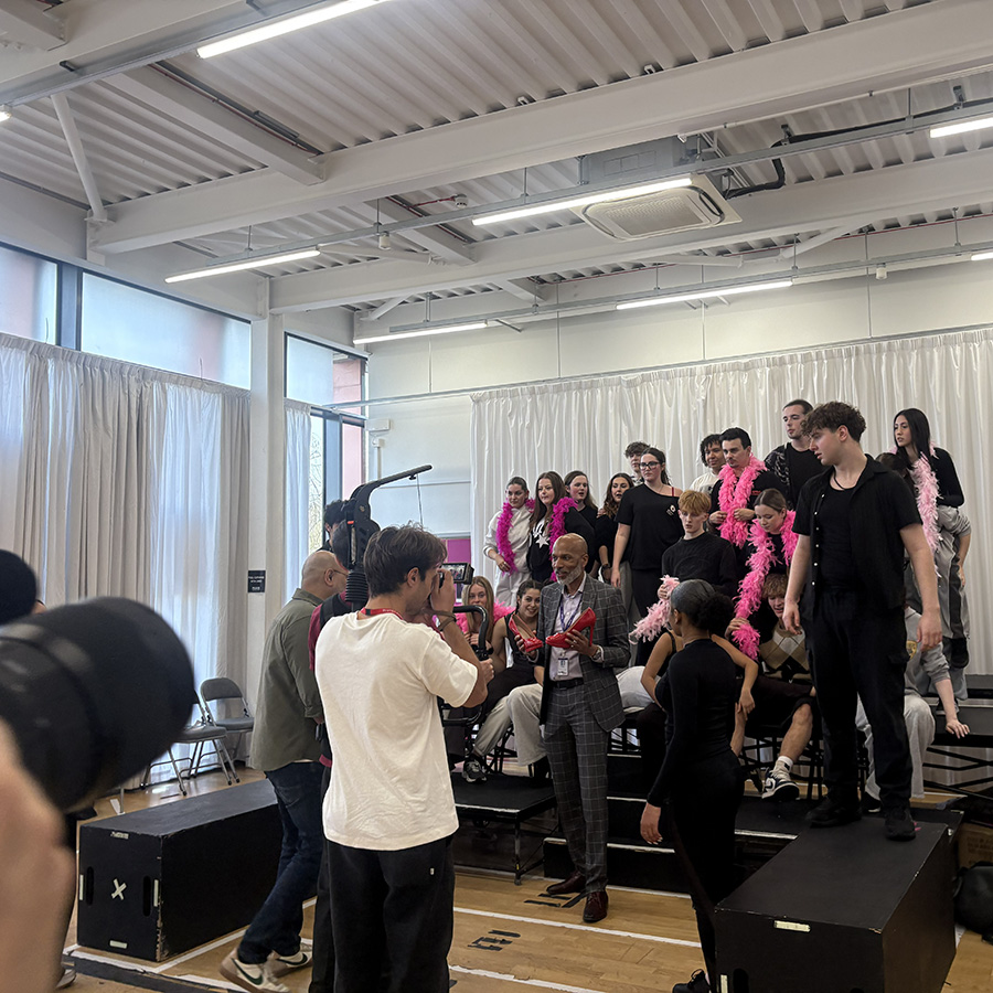 Meet the Principal Video behind the scenes footage - Clive was treated to a preview of Everybody's Talking About Jamie. In the background students are behind Clive wearing pink feather boas and he is holding red high heels in his hands. There is a film crew in the foreground. 