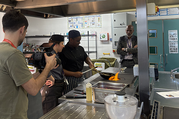 Meet the Principal Video behind the scenes footage - Clive watches Uxbridge Culinary students cooking in the Revisco kitchen - there is a flame coming out of the range stove top. A film crew is in the foreground.