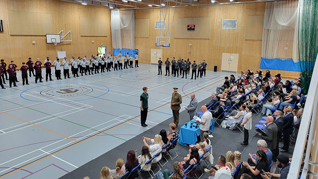 Overview of the Uniformed Services Graduation Parade at Uxbridge College in the sports hall. One student is out front receiving an award. There is an audience of family, friends and guests.