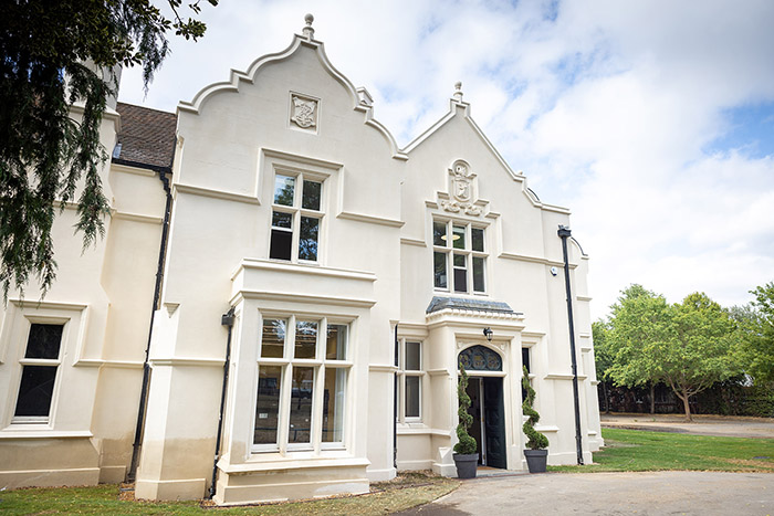 Outside of Barra Hall. White building with ornate roofing and garden area to the right with 2 plants at the entrance.