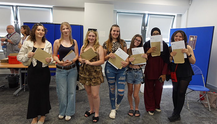 A group photo of female Uxbridge College students holding their exam results on A-Level results day. They are standing in a classroom and are all smiling.     