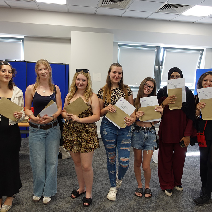 A group photo of female Uxbridge College students holding their exam results on A-Level results day. They are standing in a classroom and are all smiling.     