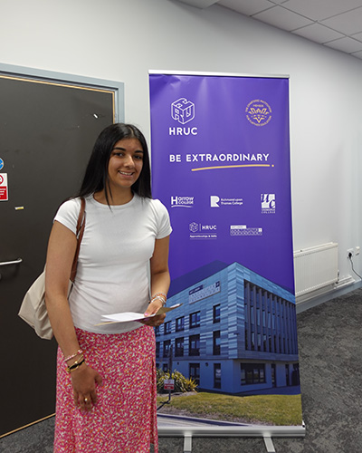 Female Uxbridge College student holding her exam results in her hand and standing in front of a Be Extraordinary banner. She looks very pleased.