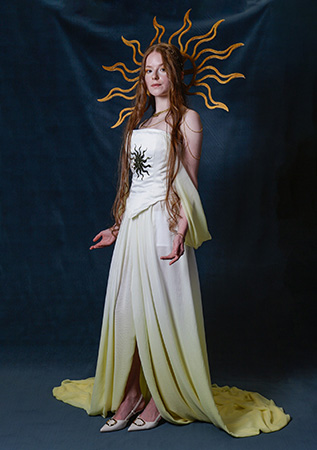 One of the Uxbridge College original designs from their fashion show. A girl with long red hair is wearing an off-white dress with a bodice that has a black sun pattern on it. She has the sun patern as a golden head piece and is holding her arms with palms up.
