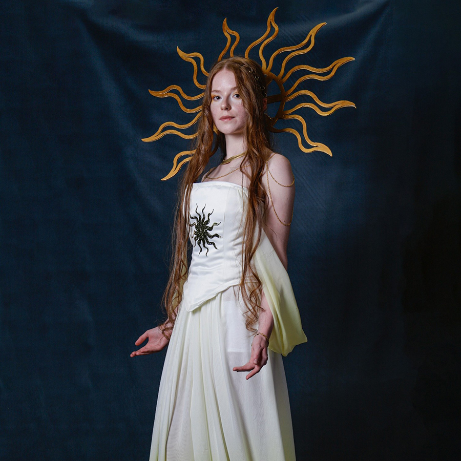 One of the Uxbridge College original designs from their fashion show. A girl with long red hair is wearing an off-white dress with a bodice that has a black sun pattern on it. She has the sun patern as a golden head piece and is holding her arms with palms up. 