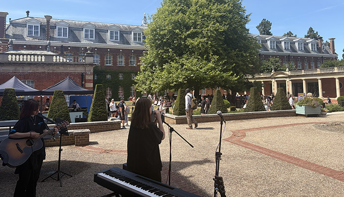 A view from behind the band looking at Wellington College and the attendees at the Festival of Education. There is a large tree in front of buildings. In the foreground there is a keyboard, a student at a microphone and one playing guitar.