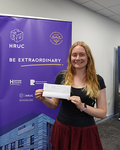 Uxbridge College student Rebecca holding her A-Level results in her hand and standing in front of a Be Extraordinary banner. She looks very happy.