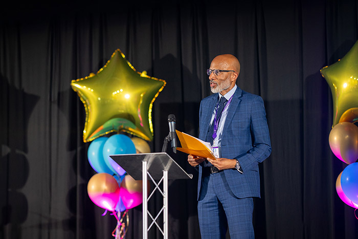 Clive Hodge, Principal of Uxbridge College, speaking at the Uxbridge College Student Awards ceremony. He is dressed in a blue check suit and has balloons at the back of him with a lectern in front of him.