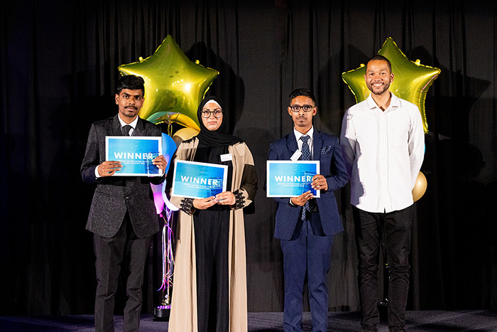 Greatest Contribution to College Life award went to three students - Saubana Zaman, Shaun Rodrigues and Larence Cardoso - standing on stage with their awards and certificates and Anthony Bennett to the right smiling. There are golden star balloons behind them with a black curtin backdrop.