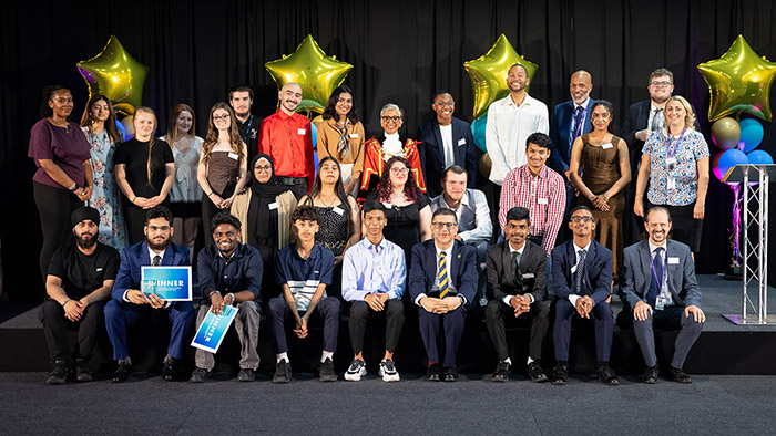 Uxbridge College Student Awards winners with staff, Clive Hodge (Principal) and Anthony Bennett. They are in a group on stage with gold star balloons behind them.