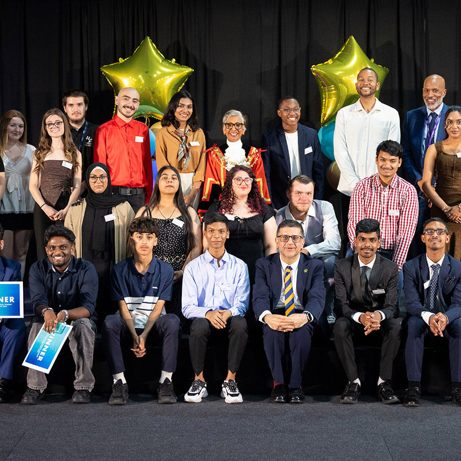 Uxbridge College Student Awards winners with staff, Clive Hodge (Principal) and Anthony Bennett. They are in a group on stage with gold star balloons behind them.