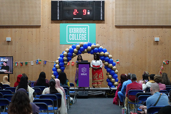 Staff speaking on stage at the Celebration Day Cultural Event. There is a gold and blue balloon arch behind them with Uxbridge College logo. Audience are sitting in front of them in blue chairs.