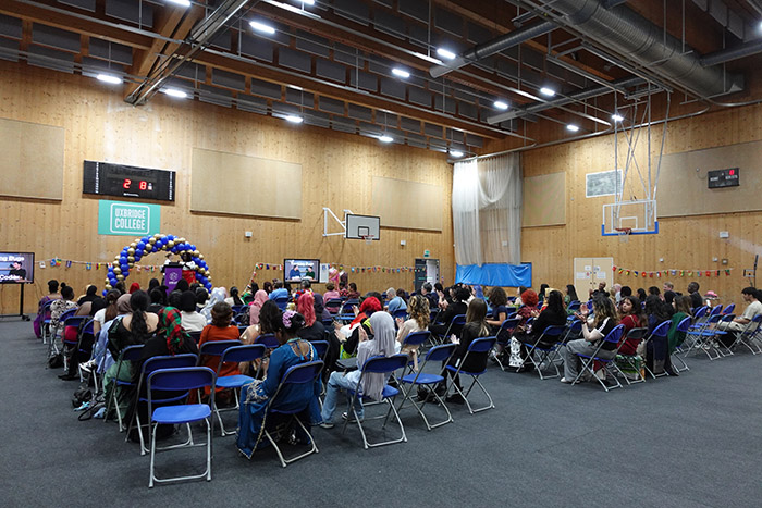 Students and staff from the Health & Social Care and Creative Arts School at the Celebration Day Cultural Event hosted in the Uxbridge College sports hall. The audience is sitting on blue chairs and there is a balloon arch at the front on a stage.