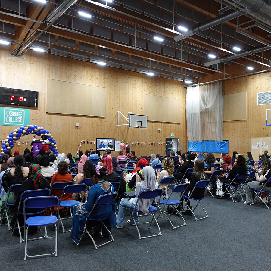 Students and staff from the Health & Social Care and Creative Arts School at the Celebration Day Cultural Event hosted in the Uxbridge College sports hall. The audience is sitting on blue chairs and there is a balloon arch at the front on a stage.