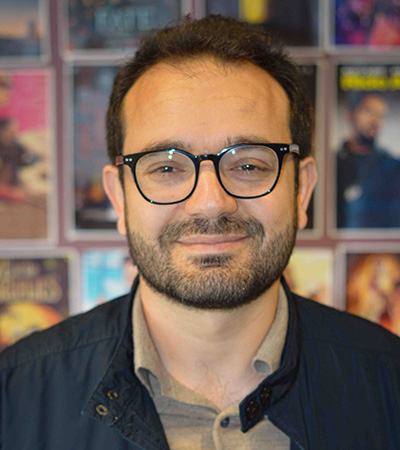 Dr Vahid Keshavarz Sarkar - he is wearing glasses, a blue jacket and khaki shirt and has a beard. Behind him is a wall of posters blurred in the background. 
