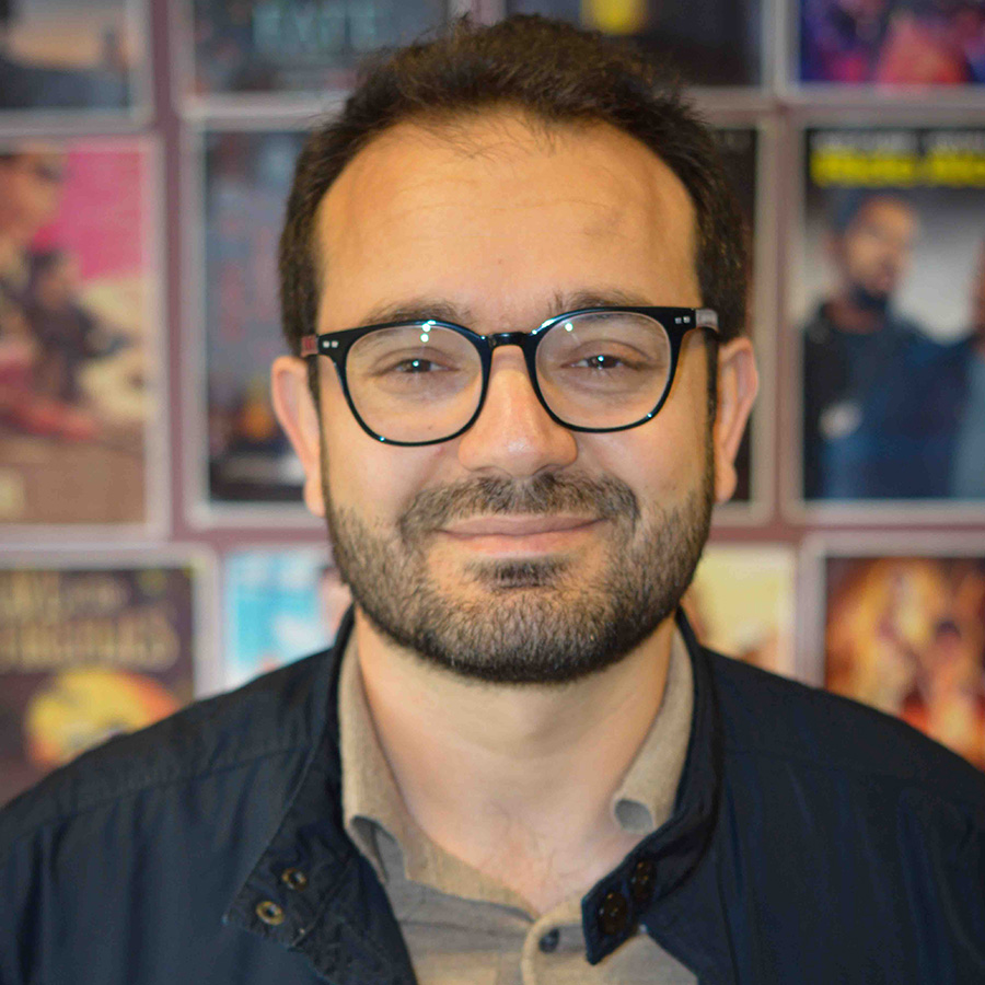 Dr Vahid Keshavarz Sarkar - he is wearing glasses, a blue jacket and khaki shirt and has a beard. Behind him is a wall of posters blurred in the background. 