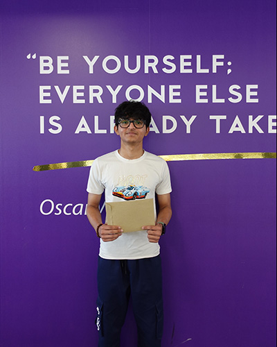 Vedant - Uxbridge College student holding his GCSE exam results envelope and standing in front of a purple wall with the quote from Oscar Wilde that says: Be yourself; everyone else is already taken. He is wearing glasses and a white t-shirt with a car on it.