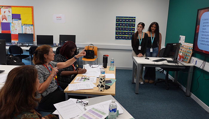 Uxbridge College staff, NHS panel and students druing the T-Level placement assessment for the Health and Social Care. Two female students are listening to the panel and standing at the front of the classroom.          
