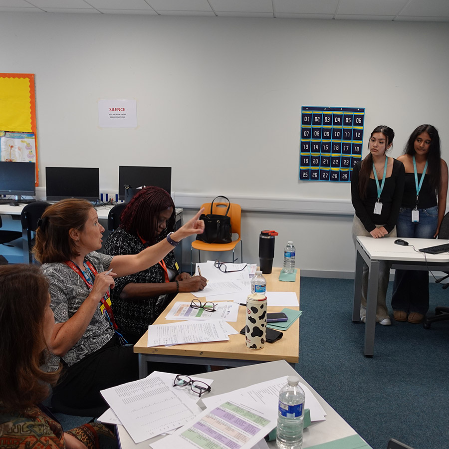 Uxbridge College staff, NHS panel and students druing the T-Level placement assessment for the Health and Social Care. Two female students are listening to the panel and standing at the front of the classroom.          